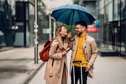 Paar steht in einer Straße und lacht. Er hält einen Regenschirm über beide, da es regnet